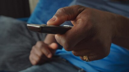 Close-up of black male hand changing tv channels with smart tv remote control while man enjoying leisure in domestic room.