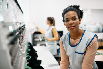 Young black woman working with embroidery machine in clothing factory and looking at camera.