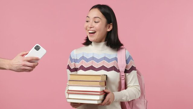 Girl teen student of Asian ethnicity wear shirt backpack hold change stack of books at mobile cell phone isolated plain light pink background studio portrait. Education in university college concept