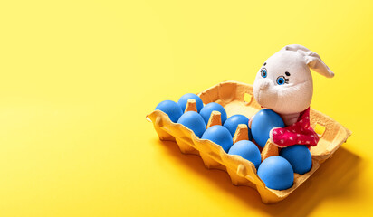 Package of ten blue painted eggs with fluffy white toy bunny on yellow background with copy space, empty text place. Banner for a religion holiday. Design mockup. Fun Easter hunt card. Handmade decor.