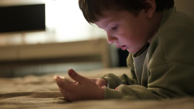 Cute little three year old child focused on smartphone late at night while lying on bed. The boy plays games on smartphone. Social and technological concept.