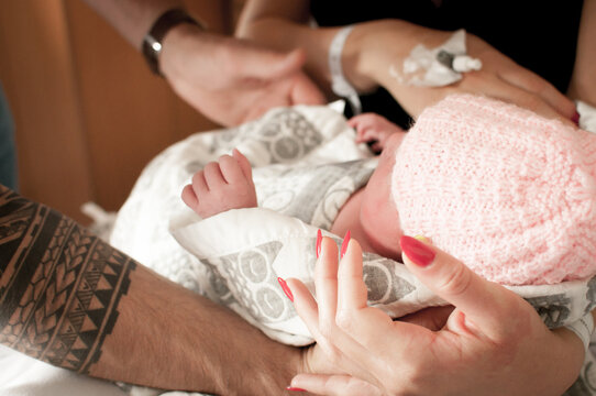 Close up of a father’s tattooed arm and her partners’ hand with red nails holding their newborn baby daughter wearing a pink hat wrapped up in a blanket in a flat in Edinburgh, Scotland, UK
