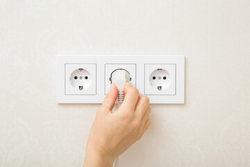 Young adult woman hand holding and plugging white electrical plug in wall outlet socket at home. Closeup. - obrazy, fototapety, plakaty