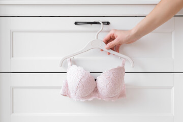 Light pink lace bra hanging on hanger at white drawer. Young adult woman hand taking underwear. Closeup. Front view. - obrazy, fototapety, plakaty