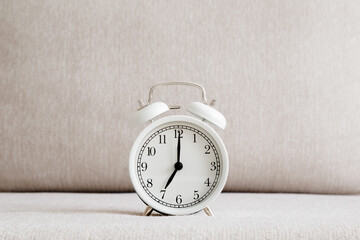 White alarm clock on light beige sofa. 7 o'clock in morning or evening. Time concept. Closeup. Front view.