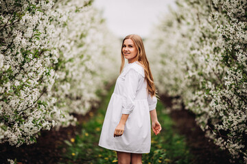 Beautiful young woman near blooming white tree in spring park. A woman in a white dress stands among the flowers on an apple tree. spring season concept