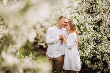 A happy young couple in love stands in a garden of blooming apple trees. A man in a white shirt and a girl in a white light dress are walking in a flowering park