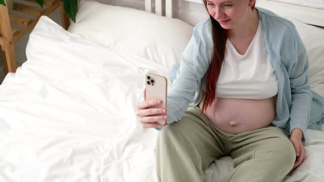 Beautiful cheerful pregnant woman taking selfie photo using mobile phone sitting on bed at home. big belly advanced pregnancy