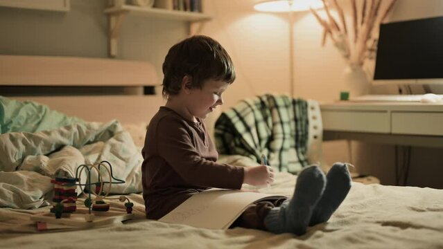 A cute little three year old child focused on drawing with pen in sketchbook late at night while sitting on the bed. The boy is learning to hold ballpoint pen and be creative.