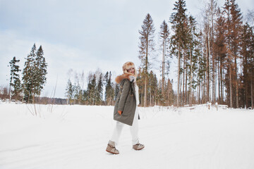 Happy smiling woman walking in winter snow nature meadow, hiking, wearing stylish fashion outfit khaki parka coat jacket, fur hat and moon boots, full length, looking back