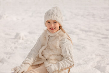 a happy little girl in a white knitted hat and sweater is sitting in a sleigh on a snow slide