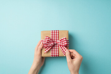 First person top view photo of valentine's day decorations girl's hands unpacking craft paper giftbox with checkered ribbon bow on isolated pastel blue background with copyspace