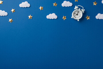 Top view photo of the small white clock with scattered golden confetti in shape of stars and white clouds on the nice deep blue isolated background copyspace