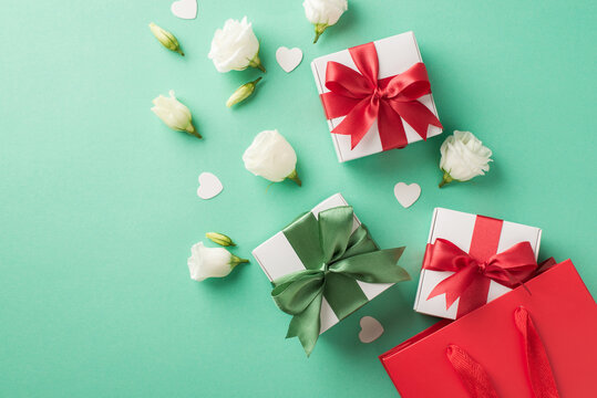 Top view photo of three small white gift boxes with cute red and green silk bows white eustomas and small confetti in shape of hearts near the big red shopping bag on the pastel turquoise background