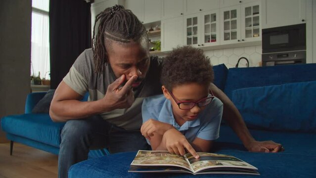 Loving caring attractive African American father and adorable elementary age son in eyeglasses talking, sharing and discussing storylines while enjoying leisure and reading comic book together.