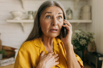 Indoor close-up image of shocked senior woman, having frightened and worried face expression after hearing bad news while talking on phone with her relatives standing against kitchen background - 484348529