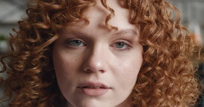 Close-up portrait of a red-haired young woman with curly hair looks at the camera and blinks her eyes, indoors at home