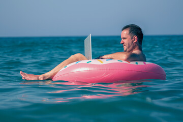 Working anywhere and any time concept.  Young man freelance businessman working at laptop on inflatable ring in the water of sea. Horizontal image.