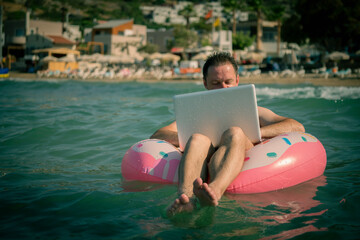 Freelancer with an inflatable ring in the water of sea works on the computer. Happy man in swimming circle with laptop. Copy space.