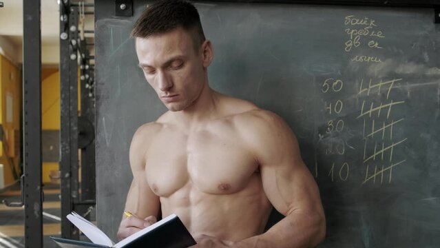 Muscular athletic man writes results in a notebook in the gym. Shoulders muscles workout. Sport, weight lifting and health care concept