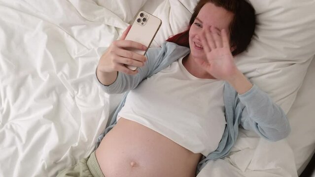 Beautiful cheerful pregnant woman taking selfie photo using mobile phone lying on bed at home. big belly advanced pregnancy