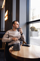 A positive man sits in a cafe at a table, he drinks aromatic coffee. Smiling guy in casual clothes sits in a cafe and drinks morning coffee