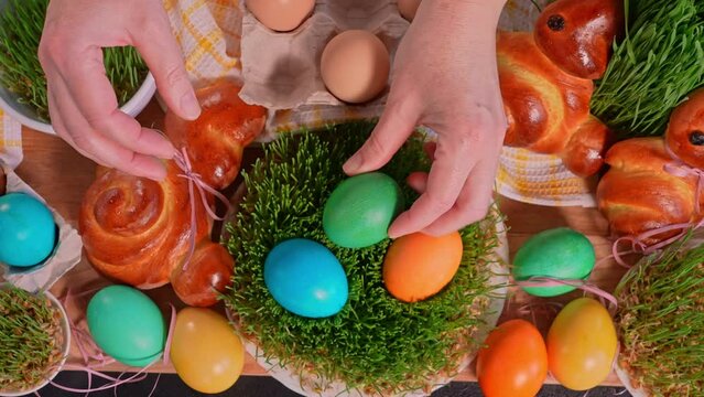 Woman`s hands laying easter eggs on freshly grown wheat sprouts, table decorated with home baked easter bunnies, top view, close-up, slow motion