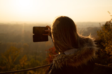 Close up photo of young woman facing away from camera, standing outdoors in the nature during autumn sunset, taking photo with her smartphone