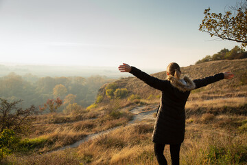 Young woman in her thirties standing with open arms in the hill area with a view on surrounding country in sunny autumn afternoon