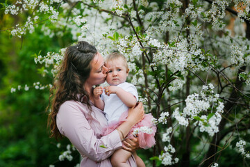 Tender mother in a pink dress holds a baby daughter in spring. Motherhood and femininity. Mom with long curly hair.