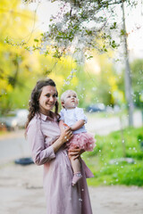 Tender mother in a pink dress holds a baby daughter in spring. Motherhood and femininity. Mom with long curly hair.