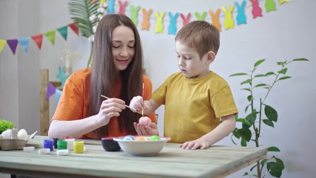 Mom and a child in bright clothes together are coloring eggs in a decorated room. Concept of family preparation for Easter, festive spring mood