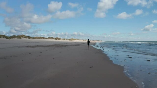 Forwards tracking of man jogging on sea coast. Sportsman running on sand beach along mild waves. Denmark