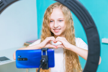 A beautiful blogger girl with long blond hair and blue eyes is dressed in a white T-shirt and shows a heart figure with her hands in honor of St. Valentine's Day and looks into a blue phone that is in