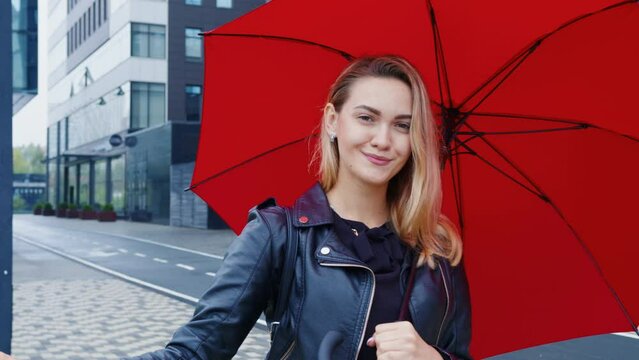 Attractive blonde in leather jacket looks at camera with loving eyes, smiles, flirts. Coquette young woman with red umbrella watches romantically at urban street. Pretty student is on date outdoor