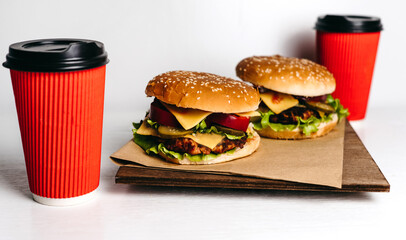 Two large burgers on kraft paper and a wooden board, with two red paper cups of coffee on a white background, a wooden table. Fast junk American food. Large portions. Takeaway delivery. Vegetarian.