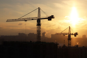 Silhouettes of construction cranes and unfinished residential building during snow on sunrise background. Housing construction, apartment block in winter city during blizzard