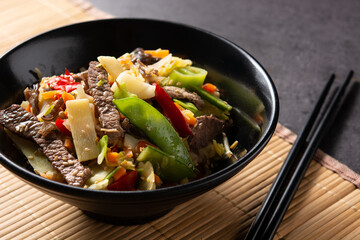 Beef, vegetables and sesame seeds in black bowl on black background