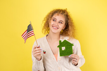 A young girl holds a model of a green house in her hands and an American flag. The concept of buying an eco house.