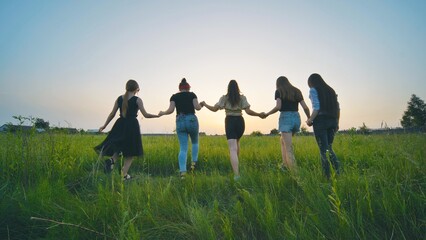Girls friends go hand in hand at sunset across the field.