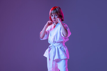 Studio shot of sportive kid, male taekwondo, karate athletes in doboks posing isolated on purple background in neon. Concept of sport, martial arts