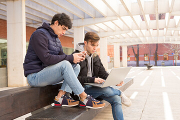 two university student friends sitting on a bench communicating with mobile phone and laptop computer