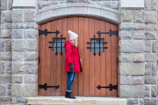 Full-length Horizontal Side View Of Pretty Young Woman With Long Blond Hair Wearing Red Puffy Winter Coat And Jeans Standing Waiting In Front Of Old Wooden Door