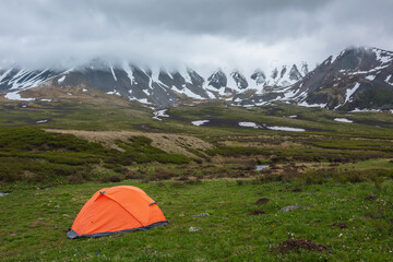 Orange tent with view to mountain range among low rainy clouds in overcast. Dramatic alpine landscape with snowy mountains in gray low clouds. Bleak atmospheric scenery of tundra under lead gray sky.