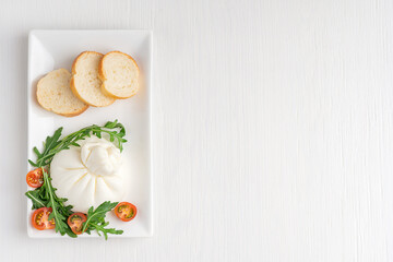 Top view of homemade organic burrata Italian milk cheese  made of mozzarella and cream served in rectangular plate with cherry tomatoes, bread and arugula on white wooden background with copy space