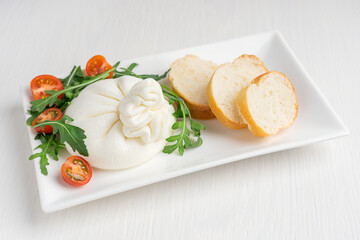 Homemade soft organic burrata Italian milk cheese made of mozzarella and cream served in rectangular plate with cherry tomatoes, arugula green leaves and sliced bread on white wooden background