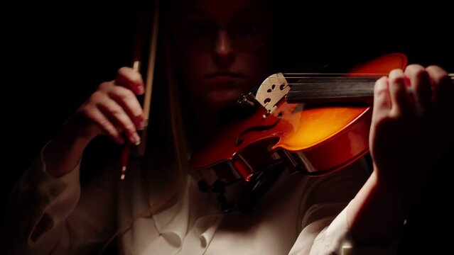 Woman with glasses bow plays the violin, standing in a dark studio. Concept of musical creativity and violin playing.