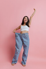 Full-length portrait of beautiful slim woman posing in huge jeans isolated over pink studio background. Weight loss concept