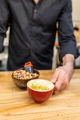 unrecognizable barista, showing a pistachio latte on the wooden bar of the coffee shop next to a bowl of pistachios.