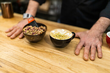 unrecognizable barista, showing a pistachio latte on the wooden bar of the coffee shop next to a bowl of pistachios.